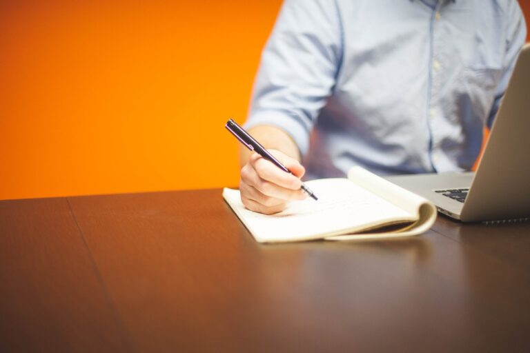 A Man Working On His Laptop And Writing Something On His Notebook