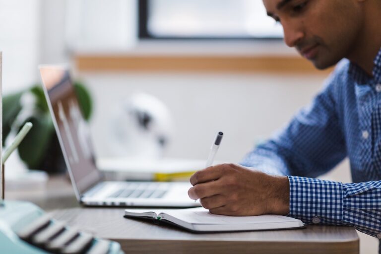 A Man Working On His Laptop And Writing Something On His Notebook